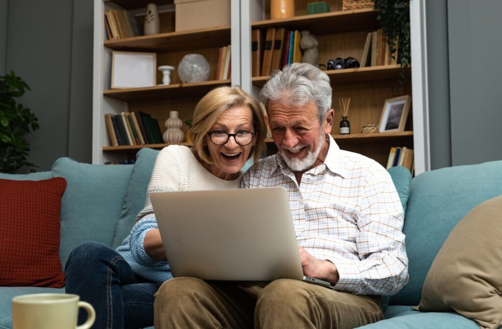 Happy senior couple are surprised by a good senior living community they are touring online, while looking at their laptop and sitting on a teal couch.