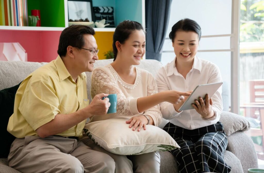 An older couple sits on a grey couch with a senior living community manager, reviewing potential lifestyles on a tablet.