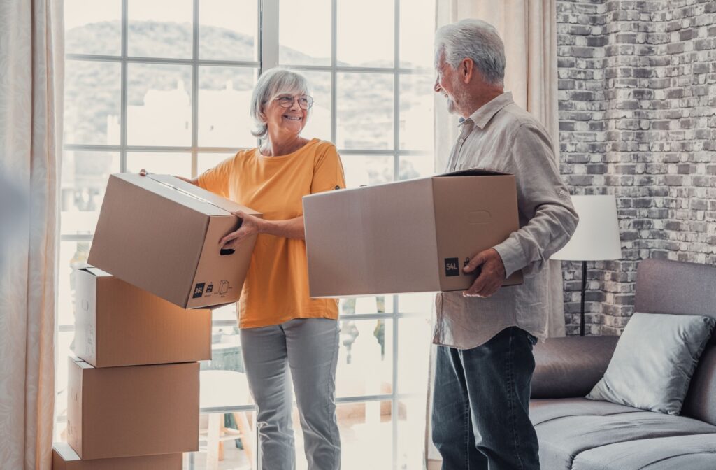 Two older adults lift boxes from in front of their window and pause to smile at one another during a move into their new home in senior living