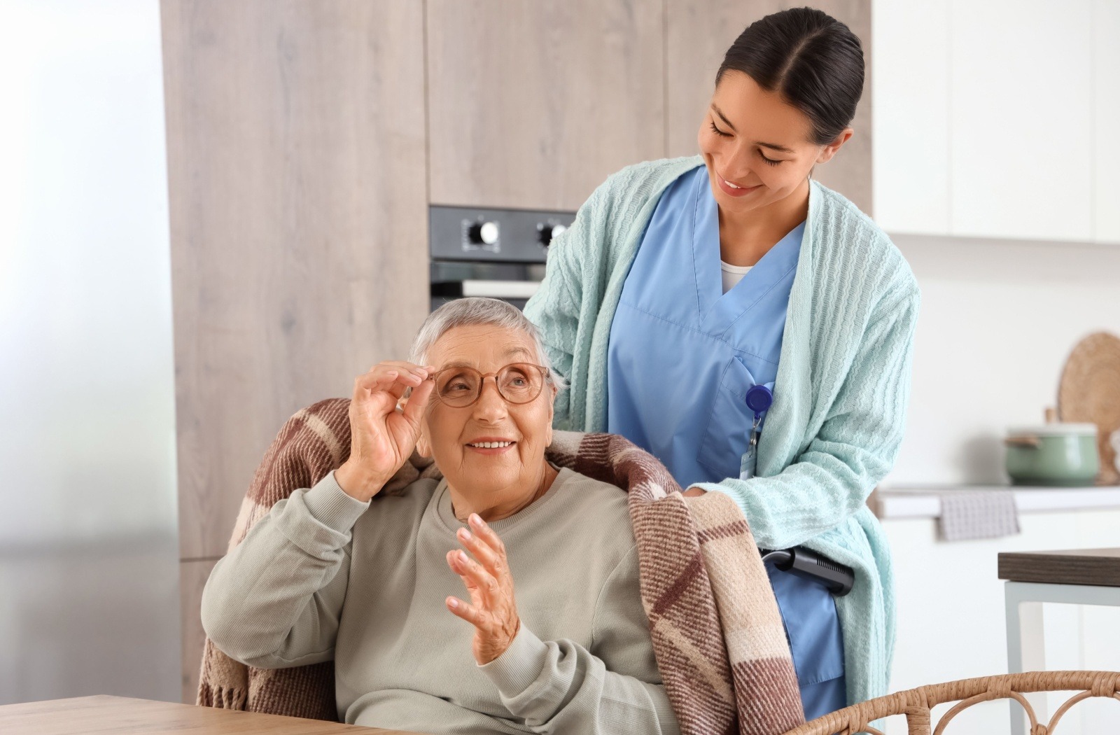 A caregiver offers a smiling older adult a cozy blanket in a bright, senior living apartment.
