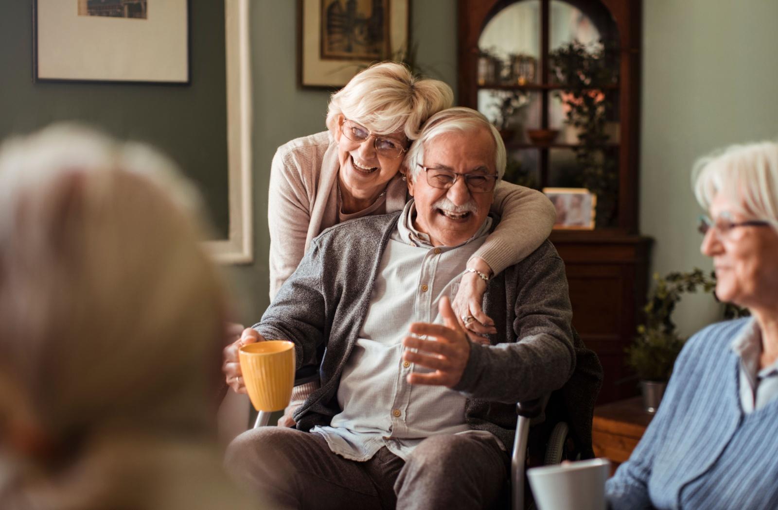 A senior couple laugh and embrace while spending time with friends they've made at their senior living community.

